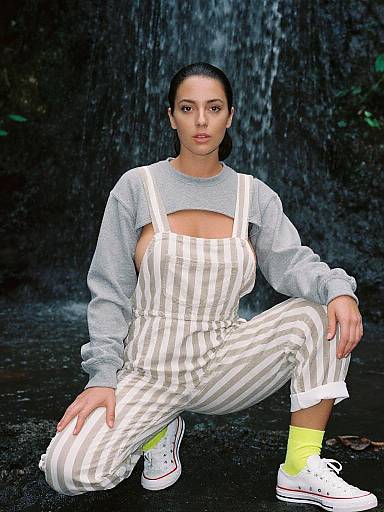 Photograph of a woman with dark hair in a bun, wearing gray long-sleeve top, white striped overalls, yellow socks, and white