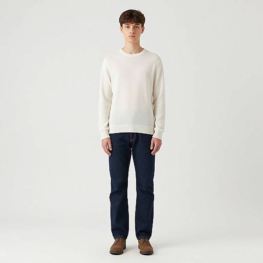 Photograph of a young man with short brown hair, wearing a white long-sleeve shirt, dark blue jeans, and brown shoes, standing against