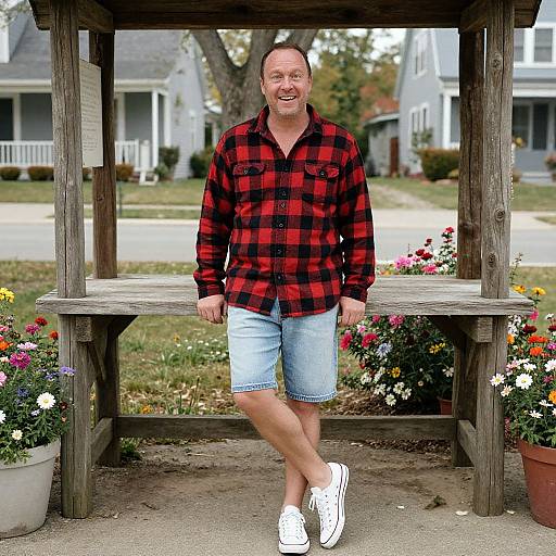 Photograph of a smiling middle-aged man in a red-black plaid shirt, light blue shorts, and white sneakers, standing under a wooden gazebo