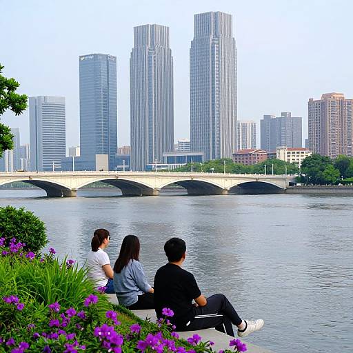 Photograph of a couple sitting by a river, facing a city skyline with tall skyscrapers and a stone bridge. Vibrant purple flowers in the