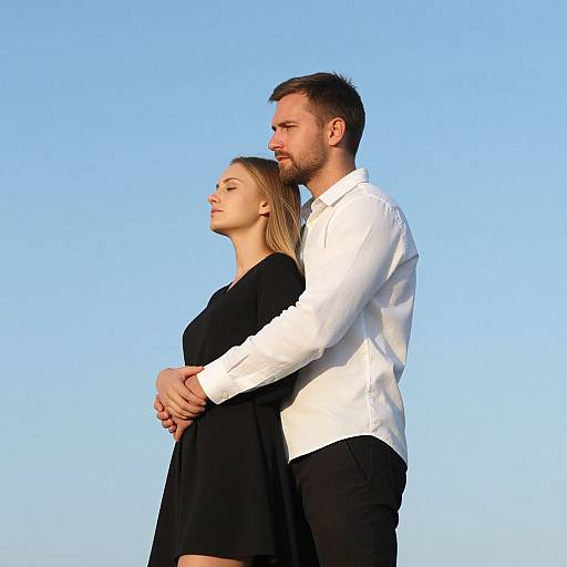 Photograph of a couple standing back-to-back, arms around each other, against a clear blue sky. He wears a white shirt, black pants;