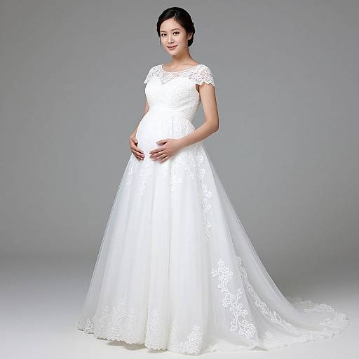 Photograph of a pregnant Asian woman in a white lace wedding dress with short sleeves and a full, flowing skirt, standing against a plain gray background.