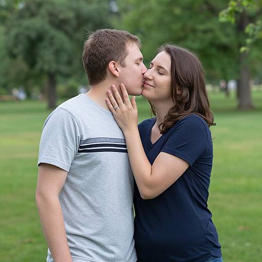 Photograph of a young couple kissing in a park, the woman in a black shirt gently touching the man's face, both smiling, surrounded by green