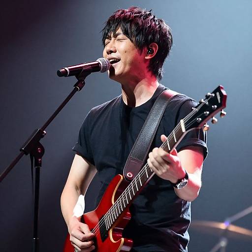 Photograph of an Asian male singer-guitarist with short black hair, wearing a black t-shirt, passionately singing into a microphone while playing a red