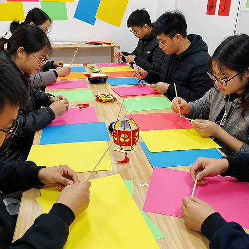 Photograph of six Asian adults, wearing black jackets, sitting at a wooden table, cutting colorful paper shapes (yellow, blue, pink), focused on