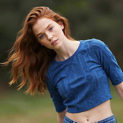 Photograph of a fair-skinned, red-haired woman with freckles, wearing a blue, patterned crop top, and jeans, leaning forward