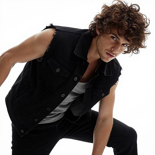 Photograph of a curly-haired, muscular man in a sleeveless black vest over a gray shirt, crouching against a white background.