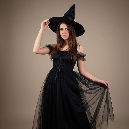 Photograph of a young woman with long brown hair, wearing a black off-shoulder witch dress and pointed hat, holding her hat and skirt against