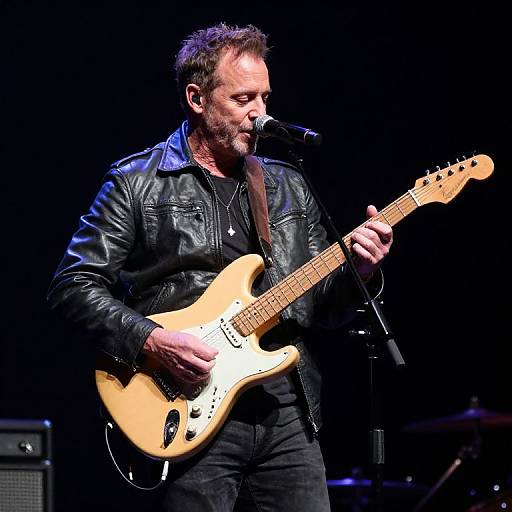 Photograph of a middle-aged man with scruffy beard, wearing black leather jacket, playing a tan electric guitar on stage, singing into microphone. Dark
