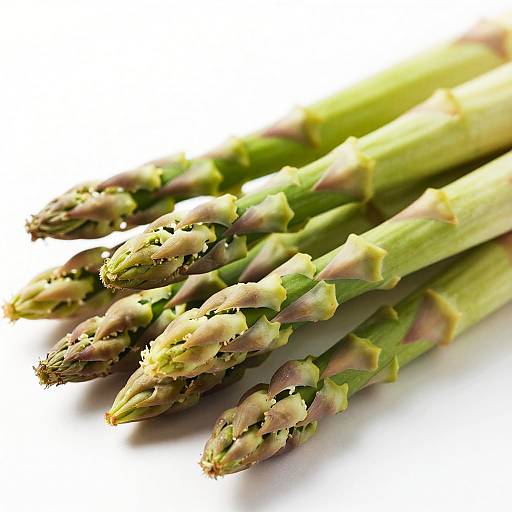 Photograph of fresh green asparagus spears with purple-tipped buds, lying horizontally on a white background, highlighting their textured, fibrous tips and