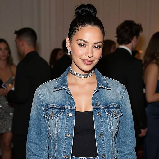 Photograph of a smiling woman with dark hair in a bun, wearing a denim jacket, black top, and sparkling choker, standing in a dim