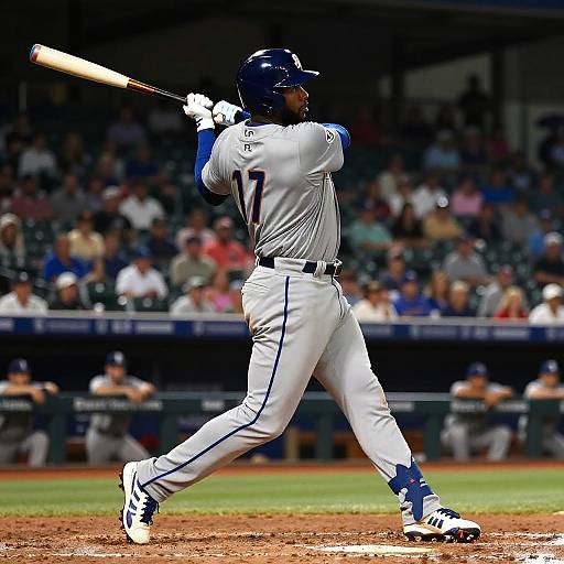 Photograph of a baseball player in white uniform with blue accents, helmet, and number 17, mid-swing in a stadium. Blurred crowd