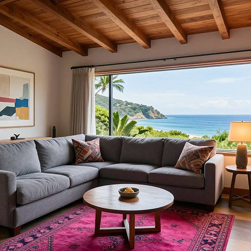 Photograph of a cozy living room with gray sectional sofa, red patterned rug, wooden ceiling beams, ocean view, and modern art.
