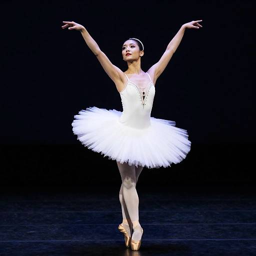 Photograph of a ballerina in a white tutu and pointe shoes, arms gracefully raised, standing en pointe against a black background.