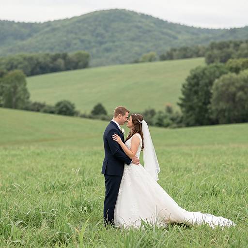 Photograph of a bride in a white dress and veil, kissing her groom in a black suit, standing in a lush green field with rolling hills and