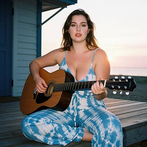 Photograph of a fair-skinned woman with shoulder-length brown hair, wearing a blue tie-dye romper, sitting on a wooden deck, playing