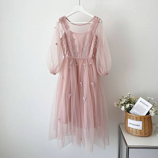 Photograph of a pink, sheer lace dress with floral embroidery, hanging on a white hanger, beside a wicker basket with white flowers and a
