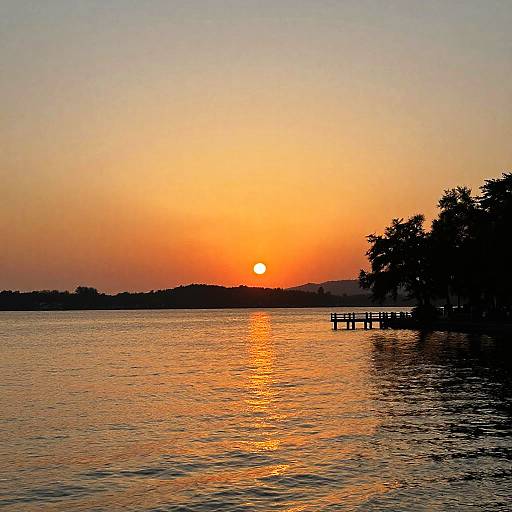 Photograph of a serene sunset over a calm lake, with a silhouetted tree on the right and a small dock extending into the water.