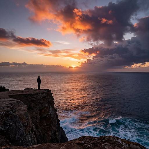 Photograph of a solitary figure standing on a rocky cliff, watching a vibrant orange and purple sunset over a vast, rippling ocean.