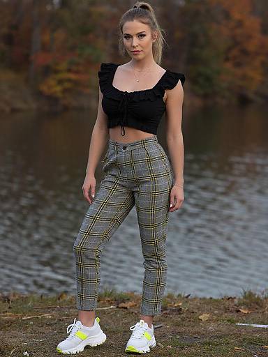 Photograph of a blonde woman with a ponytail, wearing a black ruffle crop top and plaid high-waisted pants, standing by a