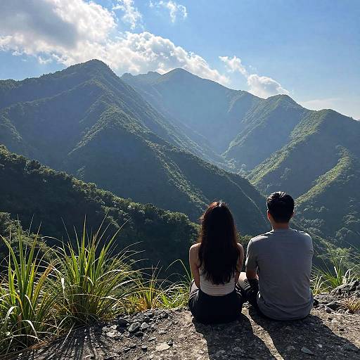 Photograph of a couple with long dark hair and short dark hair, seated on a rocky mountain ledge, overlooking lush, sunlit mountains under a partly
