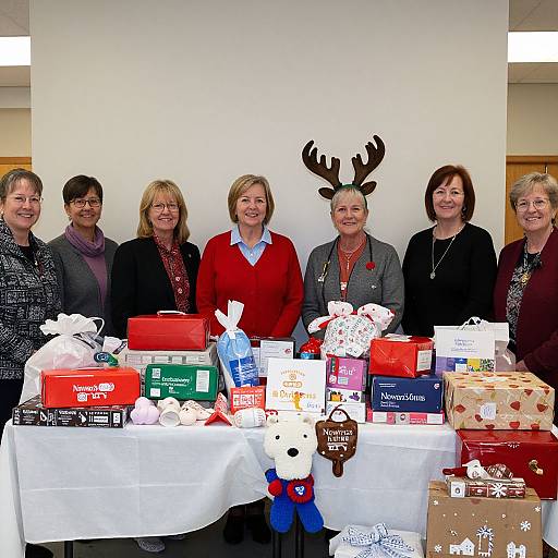 Photograph of seven middle-aged women standing behind a table filled with Christmas gifts, including boxes, cards, and a reindeer head decoration.