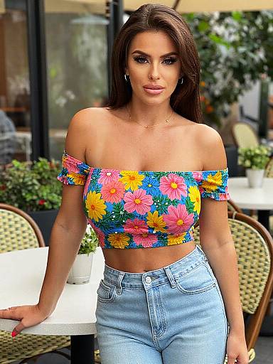 Photograph of a brunette woman with long hair, wearing a colorful off-shoulder floral top and high-waisted blue jeans, standing outdoors at