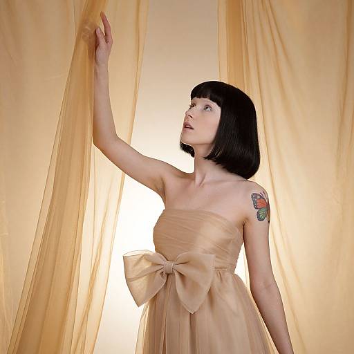 Photograph of a pale-skinned woman with black bob haircut, wearing a strapless, beige dress with large bow, touching yellow curtains, visible colorful