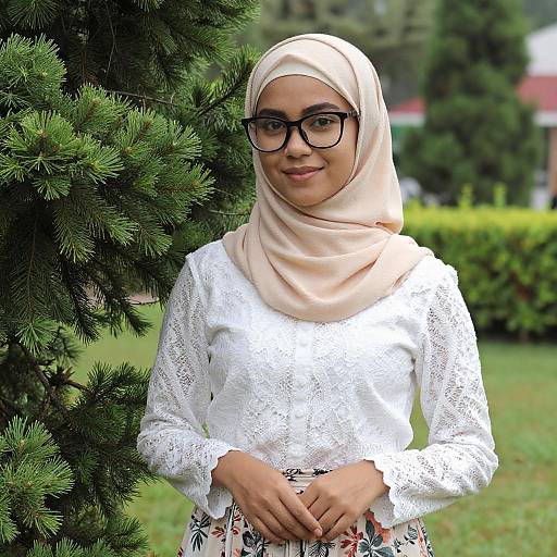 Photograph of a smiling young woman with light brown skin, black-rimmed glasses, beige hijab, white lace blouse, and floral skirt,