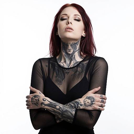 Photograph of a pale-skinned, red-haired woman with extensive black tattoos, wearing a sheer black top, arms crossed, eyes closed, white background