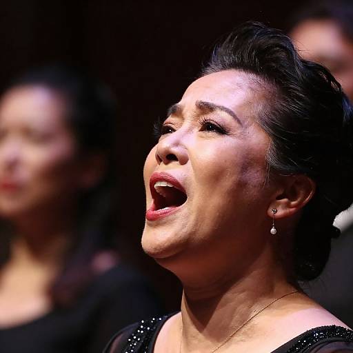 Photograph of a Latina woman with dark hair, wearing a black sequined top, passionately singing with her mouth open, illuminated by stage lights, blurred