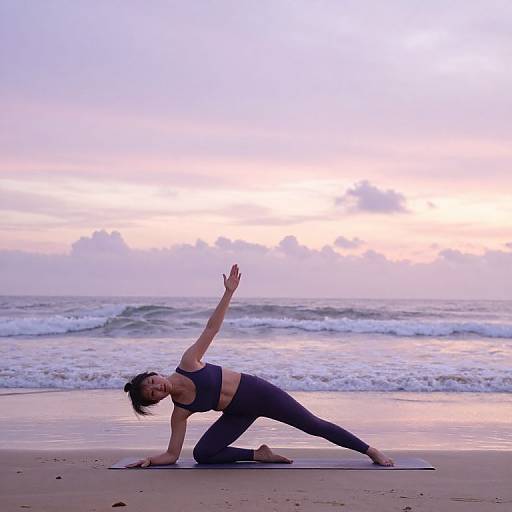Photograph of a woman in a black sports bra and leggings, performing a yoga pose on a beach at sunset. Waves and pastel sky in the
