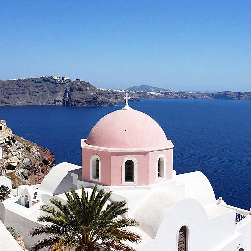 Photograph of a pink-domed church with white walls, surrounded by blue sea and palm tree, set against a rocky island landscape. Bright sunlight,