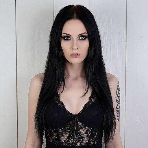 Photograph of a pale-skinned woman with long black hair, black lace top, and a tribal tattoo on her right arm, standing against a white