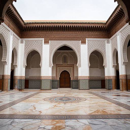 Photograph of an ornate Islamic courtyard with arched brick columns, intricate white plasterwork, patterned tiled floor, and a central wooden door.