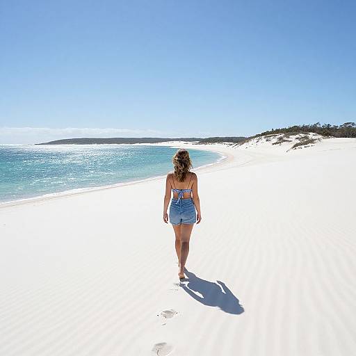Photograph of a woman with brown hair in a blue sundress walking alone on a bright white sandy beach under a clear blue sky, with turquoise ocean
