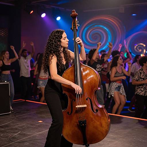 Photograph of a curly-haired woman playing a large wooden double bass on stage, with a vibrant, dancing crowd in the background under colorful, swirling lights