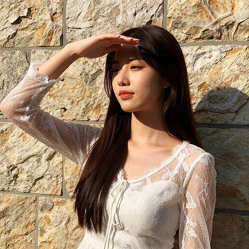 Photograph of an Asian woman with long black hair, wearing a white lace top, shading her eyes against sunlight on a textured stone wall.