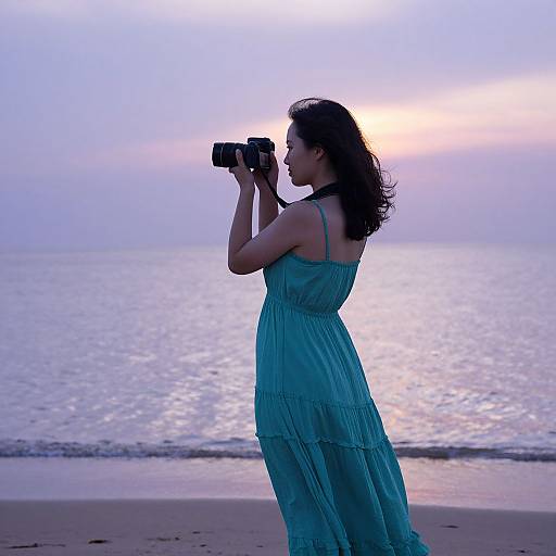 Photograph of a woman in a blue sundress, holding a camera, standing on a beach at sunset, with calm ocean waves in the background.