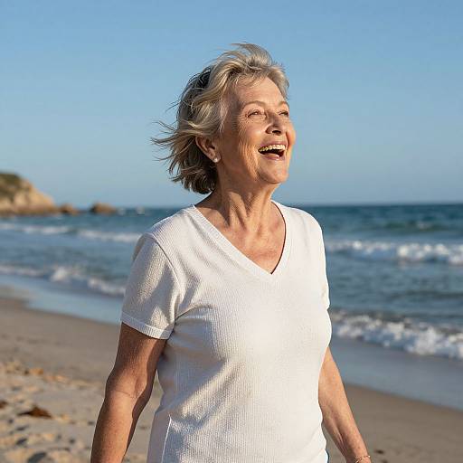 Photograph of an elderly woman with short gray hair, wearing a white shirt, smiling at a sunny beach with clear blue sky and gentle waves.