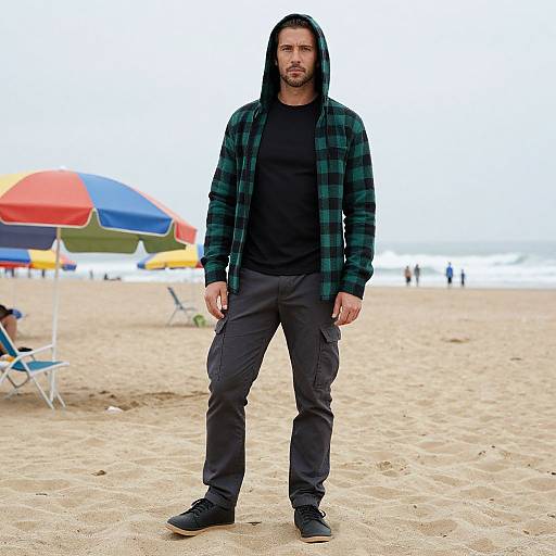 Photograph of a bearded man with long black hair, wearing a green plaid hoodie, black shirt, and dark pants, standing on a sandy