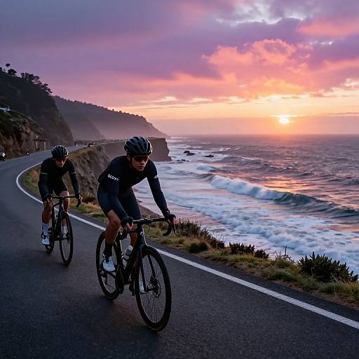 Two cyclists in black gear ride along a coastal road at sunset, with vibrant pink and orange clouds and crashing waves.
