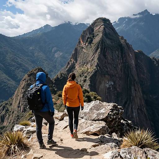 Photograph of two hikers, one in blue jacket, one in orange, climbing rocky mountain trail with towering peak and forested mountains in background under