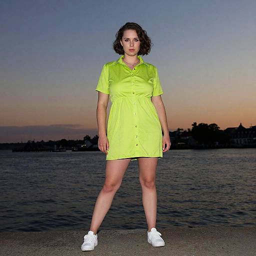 Photograph of a curly-haired woman in a neon yellow dress and white sneakers, standing confidently at dusk by a calm water body.