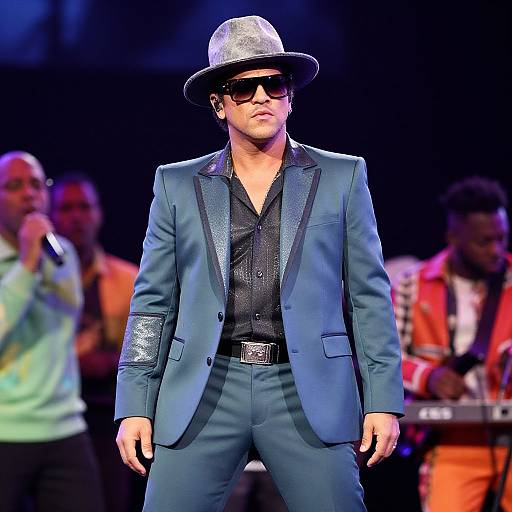 Photograph of a confident man in a blue satin suit, black shirt, silver hat, and dark sunglasses, standing on stage with blurred background musicians.