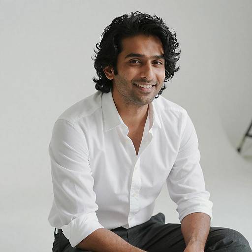 Photograph of a smiling South Asian man with medium-dark skin, black wavy hair, wearing a white button-up shirt and black pants, against a