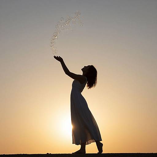 Silhouetted woman in flowing dress, arms raised, splashing water against a sunset sky. Photograph captures serene, playful moment.