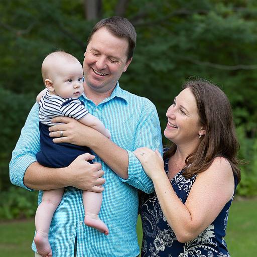 Photograph of a smiling white family outdoors: father in blue checkered shirt, holding bald baby in striped onesie, mother in black floral dress,