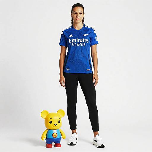 Photograph of a woman with dark hair in a blue Emirates-sponsored sports shirt, black pants, and white shoes, standing next to a yellow teddy