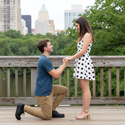Photograph of a man kneeling, holding hands with a woman in a white polka dot dress, on a wooden bridge with cityscape and greenery